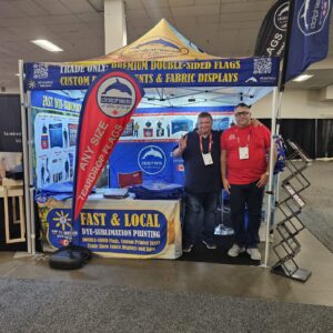 Red and blue teardrop flag on a cross base beside a trade show booth with a printed canopy tent