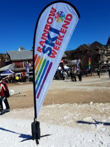 White teardrop flag with a rainbow print on a weighted base in the snow at a ski resort