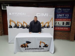 Man at a printed table in front of a white 10 ft pop up fabric display with excavator photos