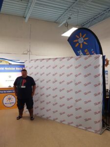 Man beside a white step-and-repeat 10 ft pop up fabric display with red logos in the workshop
