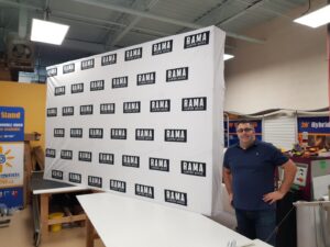 Man beside a white step-and-repeat 16 ft pop up fabric display with black logos, side view