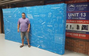 Man beside a blue 20 ft pop up fabric display with white line drawings