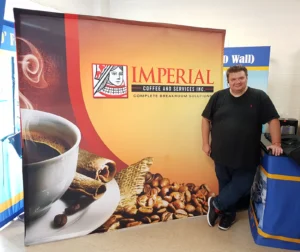 Man leaning on a counter beside an 8 ft pop up fabric display with a coffee cup photo