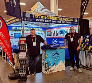 Trade show booth under a printed canopy tent with flags and a counter