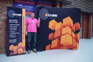 Man beside a dark blue an 8 ft curved tube fabric display with an orange cube graphic